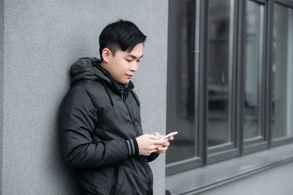 Joven asiático hombre charlando en smartphone mientras de pie por gris pared - foto de stock