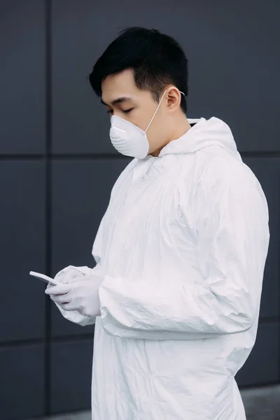 Épidémiologiste asiatique en combinaison Hazmat et masque respirateur bavarder sur smartphone tout en se tenant à l'extérieur — Photo de stock