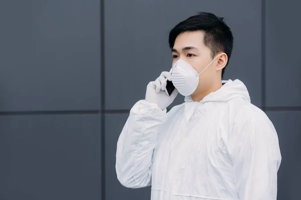Asiático epidemiólogo en hazmat traje y respirador máscara hablando en smartphone y mirando hacia fuera fuera fuera - foto de stock