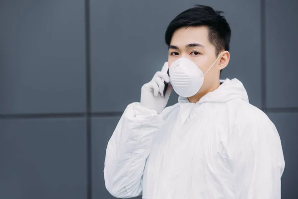 Asian epidemiologist in hazmat suit and respirator mask talking on smartphone and looking at camera while standing on street — Stock Photo