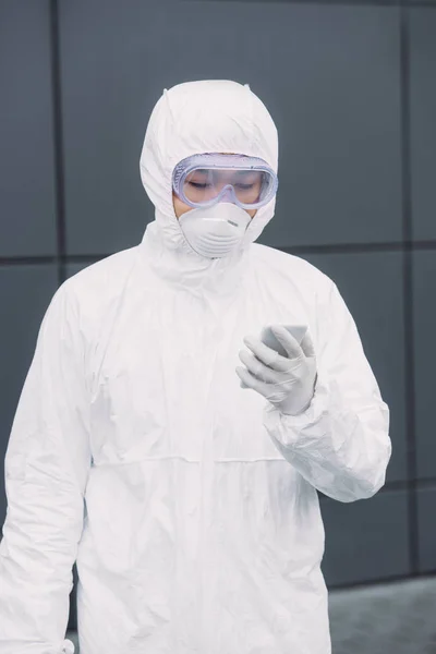 Asian epidemiologist in hazmat suit and respirator mask standing outside and looking at smartphone — Stock Photo