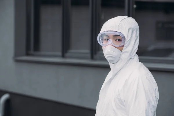 Épidémiologiste asiatique en combinaison Hazmat et masque respirateur regardant la caméra tout en se tenant debout sur la rue — Photo de stock