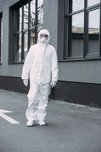 Asian epidemiologist in hazmat suit and respirator mask looking at camera while standing on street near building — Stock Photo
