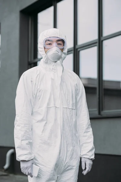 Asiatischer Epidemiologe in Schutzanzug und Atemschutzmaske schaut weg, während er in der Nähe des Gebäudes steht — Stockfoto