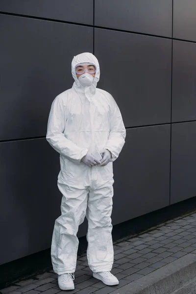 Азиатский эпидемиолог в защитном костюме и респираторной маске, смотрящий в камеру, стоя у стены — стоковое фото
