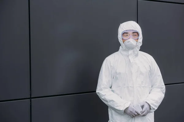 Asiatischer Epidemiologe in Warnanzug und Atemschutzmaske blickt in die Kamera, während er an der Wand steht — Stockfoto