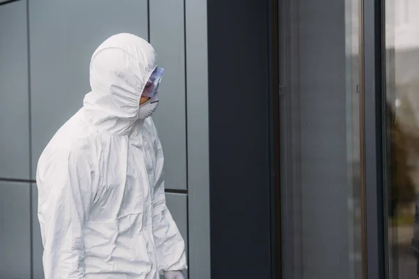 Epidemiologist in hazmat suit standing outside and looking in window of building — Stock Photo