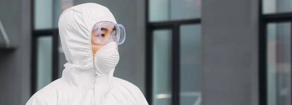 Panoramic shot of asian epidemiologist in hazmat suit and respirator mask looking away while standing on street near building — Stock Photo