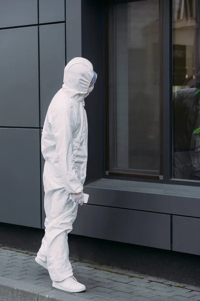 Epidemiologist in hazmat suit standing on street and looking in window of building — Stock Photo