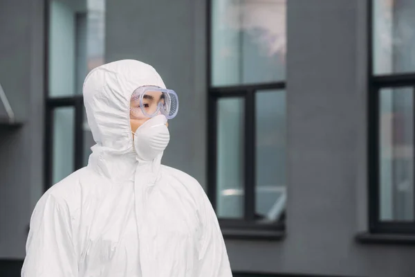 Азиатский эпидемиолог в защитном костюме и респираторной маске смотрит в сторону, стоя рядом со зданием — стоковое фото
