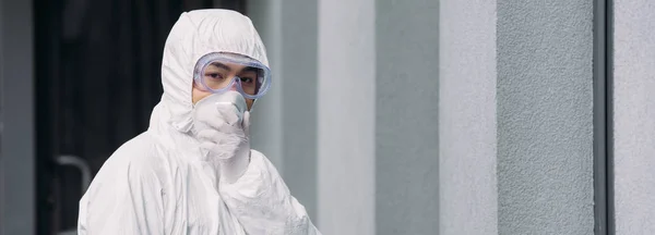 Panoramic shot of asian epidemiologist in hazmat suit and respirator mask looking at camera while standing outside — Stock Photo