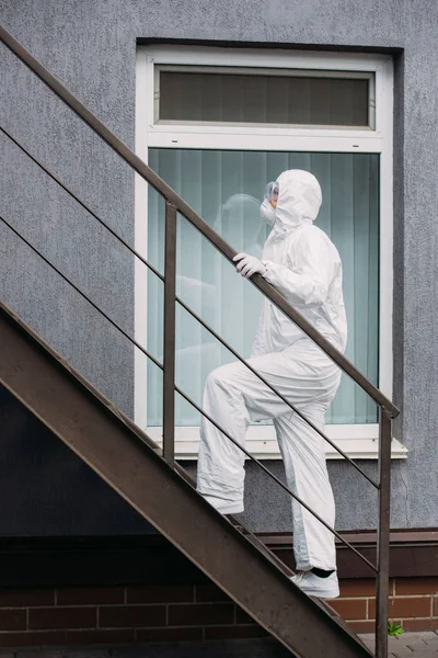 Asiatischer Epidemiologe in Warnanzug und Atemschutzmaske, der vor dem Gebäude die Treppe hochgeht — Stockfoto