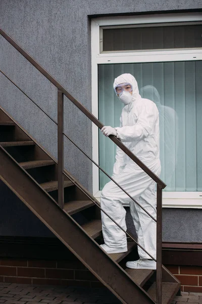 Asian epidemiologist in hazmat suit and respirator mask walking upstairs outside building — Stock Photo
