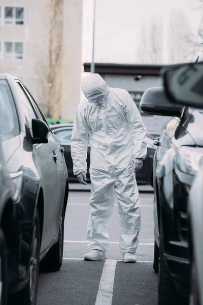 Enfoque selectivo de epidemiólogo asiático en traje de materiales peligrosos y máscara respiratoria inspeccionar vehículos en el estacionamiento - foto de stock