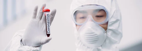 Panoramic shot of asian epidemiologist in hazmat suit and respirator mask looking at test tube with blood sample while standing outdoors — Stock Photo