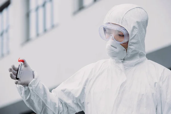 Asiático epidemiologista em hazmat terno e respirador máscara segurando tubo de teste com amostra de sangue enquanto em pé ao ar livre — Fotografia de Stock