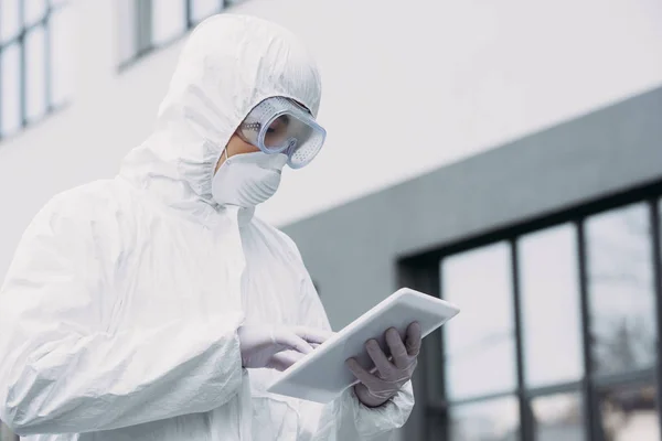 Épidémiologiste asiatique en combinaison Hazmat et masque respirateur à l'aide d'une tablette numérique tout en se tenant debout sur la rue — Photo de stock