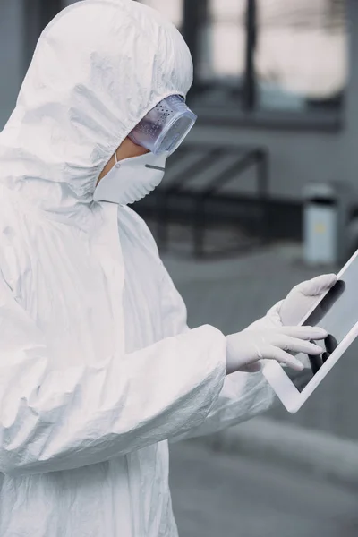 Epidemiólogo asiático en traje de hazmat y máscara respiratoria usando tableta digital mientras está de pie en la calle - foto de stock
