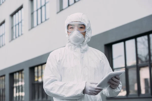 Asiatischer Epidemiologe in Warnanzug und Atemmaske mit digitalem Tablet und Blick in die Kamera, während er auf der Straße steht — Stockfoto