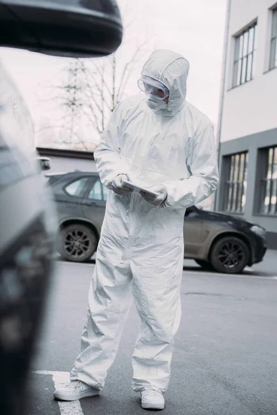 Enfoque selectivo del epidemiólogo en traje de materiales peligrosos que sostiene la tableta digital mientras revisa los coches en el estacionamiento - foto de stock