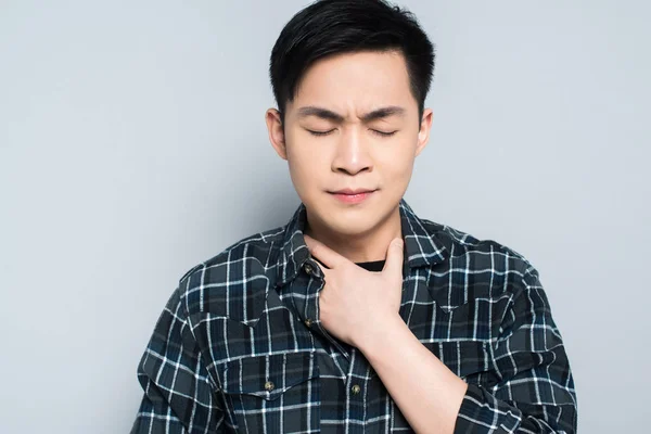 Junger asiatischer Mann mit geschlossenen Augen, der Hals berührt, während er an Halsschmerzen leidet, isoliert auf grau — Stockfoto