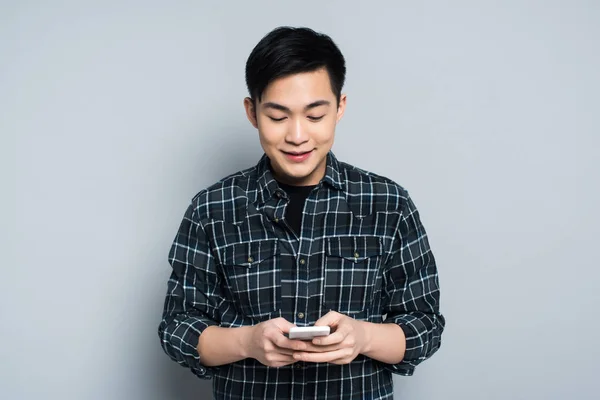Молодой азиат улыбается во время использования смартфона на сером фоне — стоковое фото