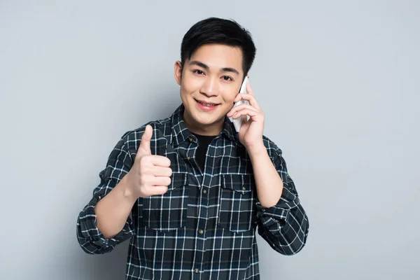 Молодий азіатський чоловік посміхається на камеру під час розмови на смартфоні і показує великий палець на сірому фоні — стокове фото