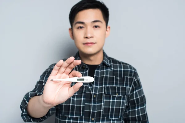 Selective focus of young asian man holding thermometer while looking at camera on grey background — Stock Photo