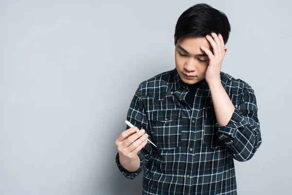 Junger asiatischer Mann blickt auf Thermometer und berührt Stirn auf grauem Hintergrund — Stockfoto