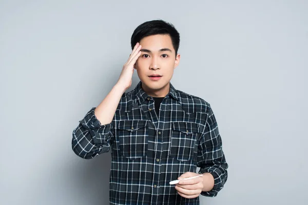 Shocked asian man touching forehead and looking at camera while holding thermometer on grey background — Stock Photo