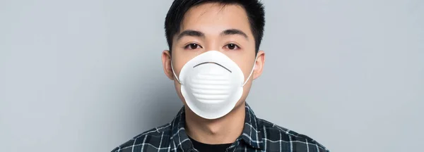 Panoramic shot of young asian man in respirator mask looking at camera on grey background — Stock Photo