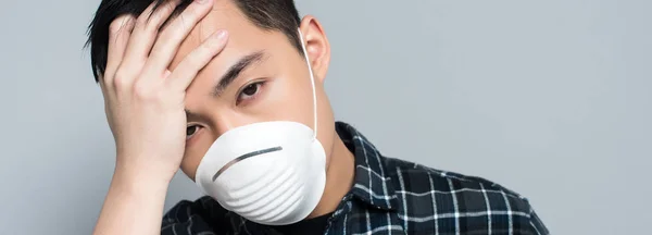 Panoramaaufnahme eines jungen asiatischen Mannes mit Atemschutzmaske, der die Stirn berührt und in die Kamera schaut, während er an Kopfschmerzen leidet, isoliert auf grau — Stockfoto