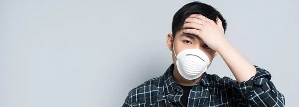 Panoramaaufnahme eines jungen asiatischen Mannes mit Atemschutzmaske, der die Stirn berührt und in die Kamera schaut, während er an Kopfschmerzen leidet, isoliert auf grau — Stockfoto
