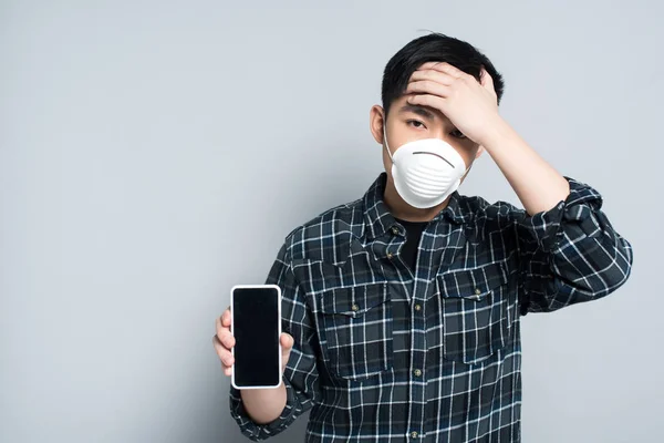 Young asian man in respirator mask touching forehead while showing smartphone with blank screen on grey background — Stock Photo