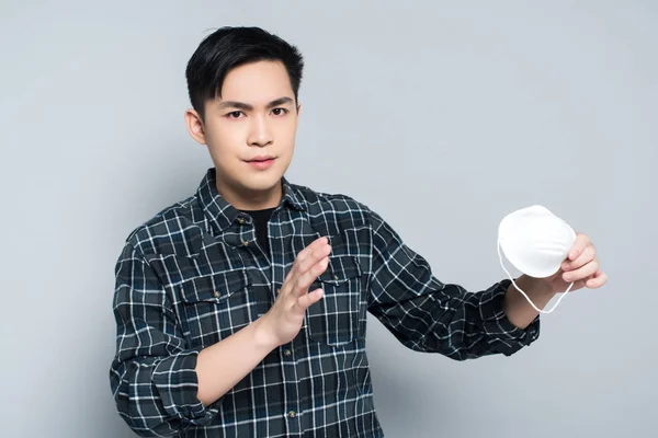 Joven asiático hombre mostrando basura gesto mientras celebración respirador máscara y mirando cámara en gris fondo - foto de stock