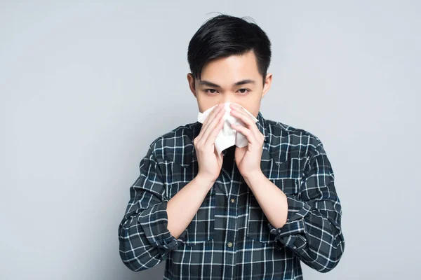 Молодий азіатський чоловік витирає ніс паперовою серветкою, страждаючи від нежить на сірому фоні — Stock Photo
