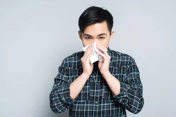 Young asian man wiping nose with paper napkin while suffering from runny nose on grey background — Stock Photo