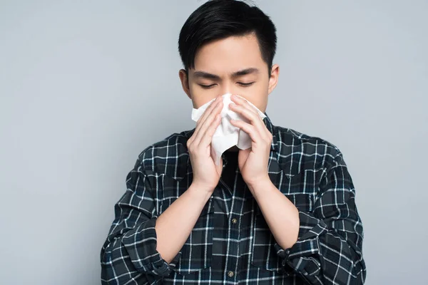 Joven asiático hombre con cerrado ojos limpiando nariz con papel servilleta mientras padece de secreción nasal aislado en gris - foto de stock
