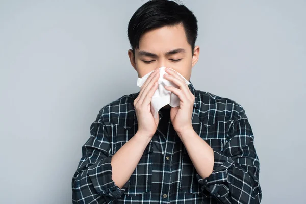 Young asian man with closed eyes wiping nose with paper napkin while suffering from runny nose isolated on grey — Stock Photo