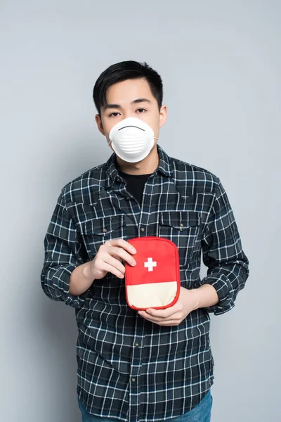 Young asian man in respirator mask showing first aid kit while looking at camera on grey background — Stock Photo