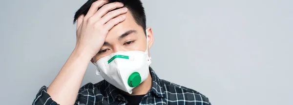 Plan panoramique de jeune homme asiatique en masque respiratoire touchant le front tout en souffrant de maux de tête isolé sur gris — Photo de stock