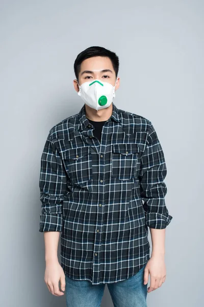 Junger asiatischer Mann mit Atemschutzmaske blickt vor grauem Hintergrund in die Kamera — Stockfoto