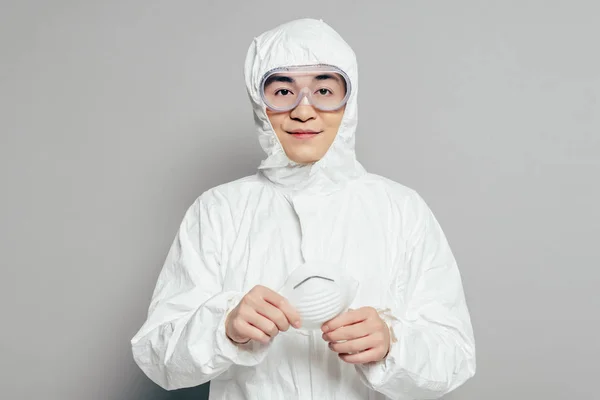 Épidémiologiste asiatique en combinaison Hazmat tenant masque respirateur tout en regardant la caméra sur fond gris — Photo de stock