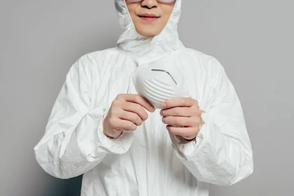 Обрезанный вид эпидемиолога в защитном костюме, держащего респираторную маску на сером фоне — стоковое фото