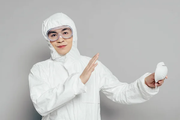 Ásia epidemiologista no hazmat terno mostrando nenhum gesto enquanto segurando respirador máscara e olhando para câmera no cinza fundo — Fotografia de Stock