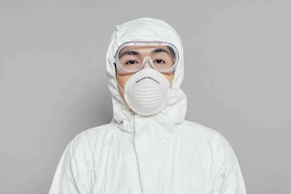 Азіатський епідеміолог у косметичному костюмі та масці для респіраторів дивиться на камеру на сірому фоні — стокове фото