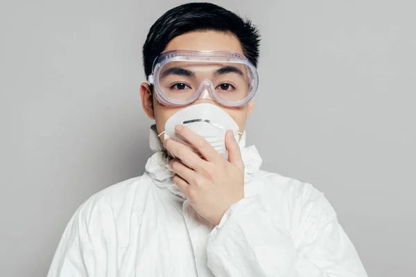 Азіатський епідеміолог у косметичному костюмі, торкаючись маски для респіраторів, дивлячись на камеру ізольовано на сірому — стокове фото