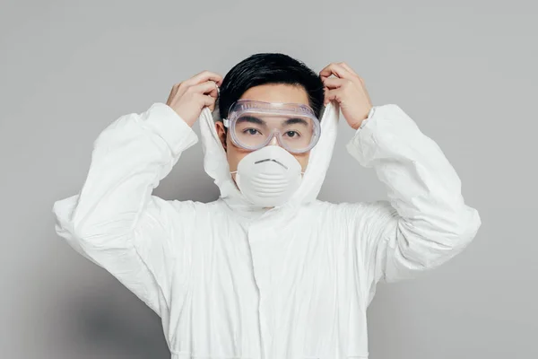 Asian epidemiologist in hazmat suit and respirator mask putting hood on while looking at camera on grey background — Stock Photo