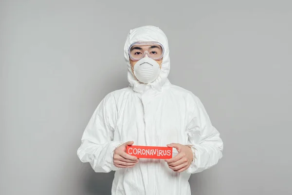Asian epidemiologist in hazmat suit and respirator mask holding warning card with coronavirus inscription on grey background — Stock Photo