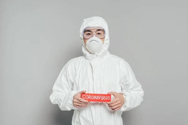 Asiático epidemiologista em terno hazmat e máscara respirador segurando cartão de advertência com inscrição coronavírus em fundo cinza — Fotografia de Stock